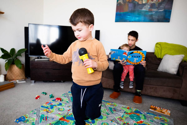 A parent is reading one child a book on the couch, while another child plays independently with a toy microphone.