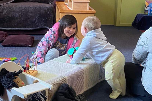 Te Ataraupo showing aroha to a another child much younger than her by talking to them over a low table.